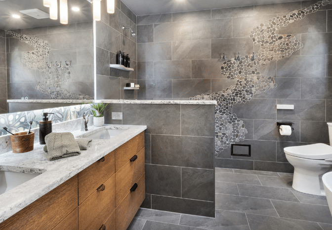 Modern bathroom featuring gray porcelain tile, a custom river rock mosaic design in the shower and wall, a dual-sink vanity with wood cabinetry and marble countertop, and contemporary lighting above a wide mirror.