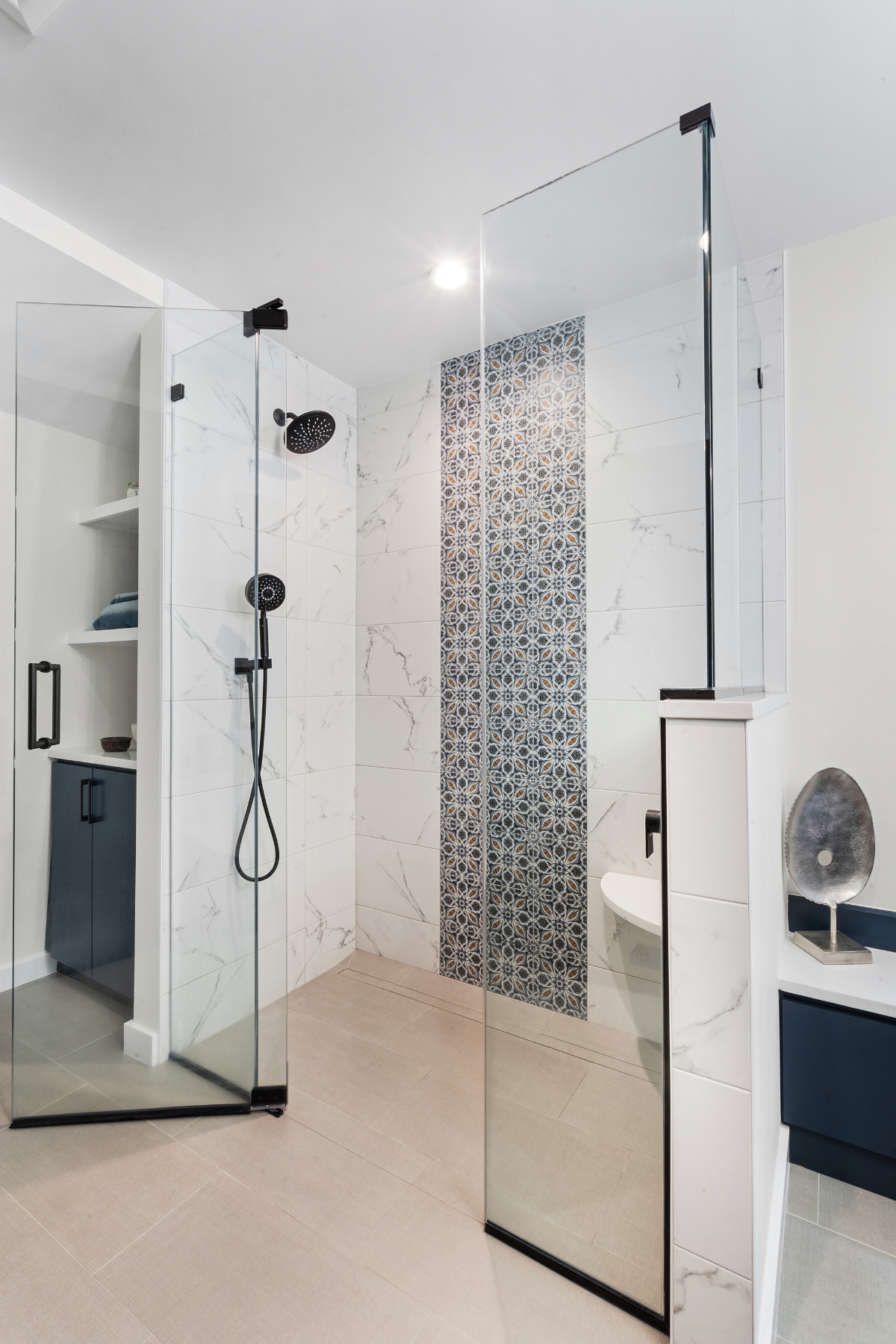 Curbless walk-in shower with marble-look tile walls, a decorative vertical mosaic accent, matte black fixtures, and glass enclosure, located in a modern bathroom with beige floor tiles and navy blue cabinetry. Curbless walk-in shower with marble-look tile walls, a decorative vertical mosaic accent, matte black fixtures, and glass enclosure, located in a modern bathroom with beige floor tiles and navy blue cabinetry.