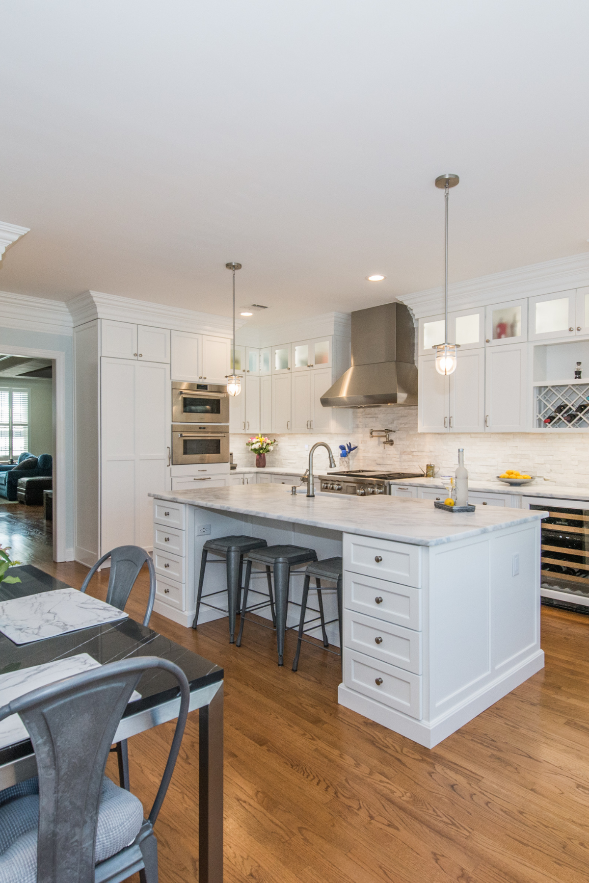 Modern white kitchen with marble countertops, stainless steel appliances, wine storage, and large center island with seating, hardwood floors, and pendant lighting.