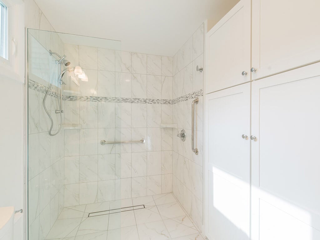 White handicap bathroom remodel with mosaic tile with accent stripe in roll-in shower with linear shower drain, grab bar, handheld and glass panel with white linen cabinet in Mount Tabor, NJ renovated by JMC Home Improvement Specialists
