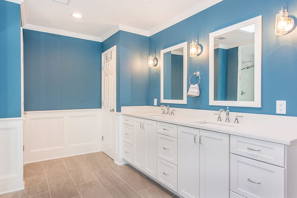 Master bathroom remodel with white wall paneling, blue painted, wood like tile floor, white shaker his and hers vanity with quartz counters and white framed mirrors with sconces in Chester, NJ renovated by JMC Home Improvement Specialists