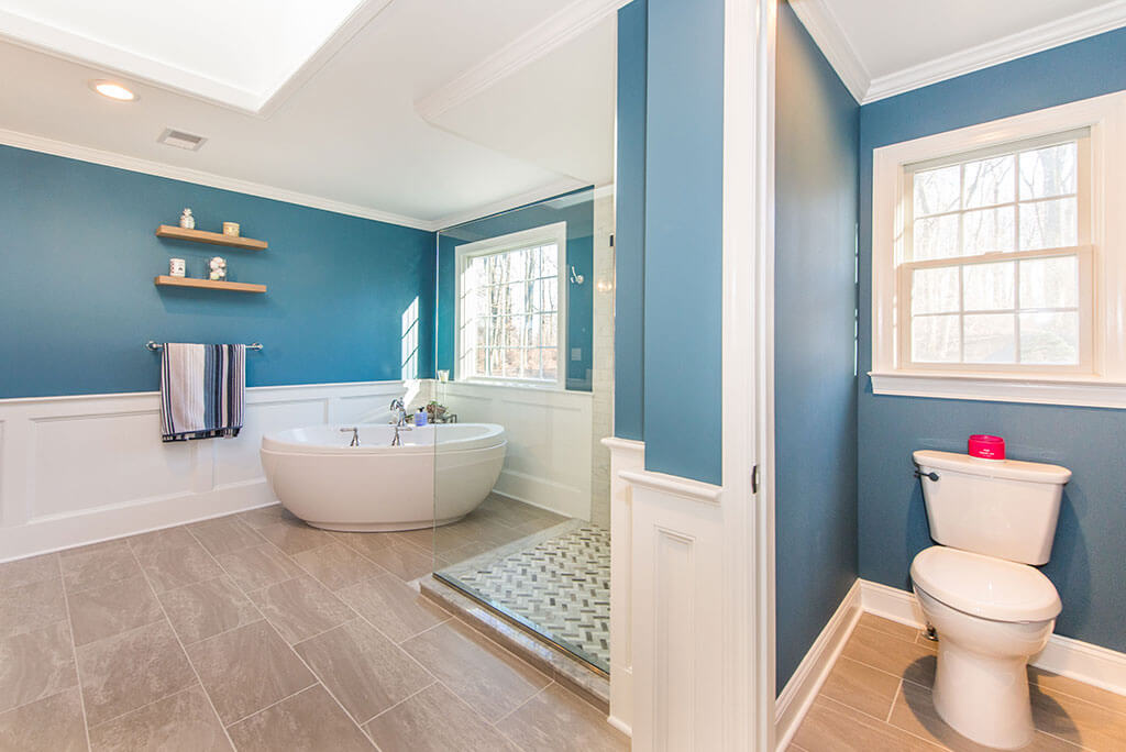Master bathroom remodel with white wall paneling, blue painted walls, clear glass shower panel, freestanding soaking tub, wood like tile floor and toilet room in Chester, NJ renovated by JMC Home Improvement Specialists