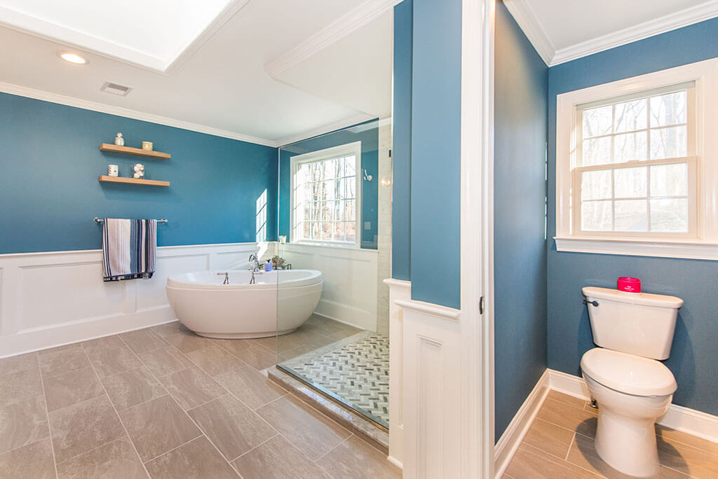Master bathroom remodel with white wall paneling, blue painted walls, clear glass shower panel, freestanding soaking tub, wood like tile floor and toilet room in Chester, NJ renovated by JMC Home Improvement Specialists
