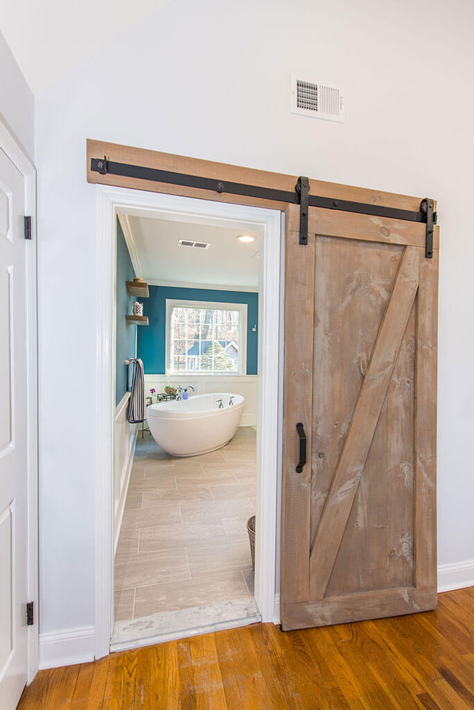 Barn door to master bathroom remodel with white wall paneling, blue painted walls, freestanding soaking tub and wood like tile floor in Chester, NJ renovated by JMC Home Improvement Specialists