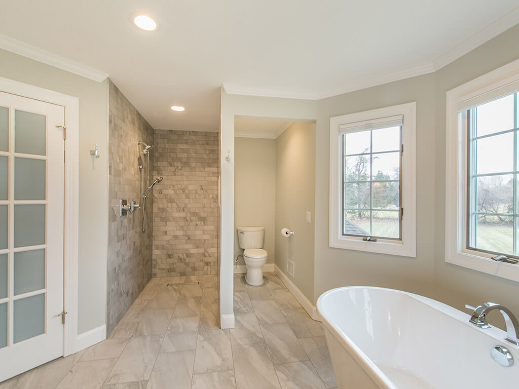Elegant master bathroom remodel with large floor tile, roll-in shower with linear shower drain, toilet room and soaking tub surrounded by Andersen windows with white trim, LED highhats and French doors in Morris County, NJ renovated by JMC Home Improvement Specialist