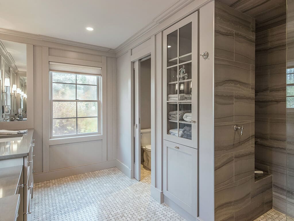 Elegant monochromatic taupe master bathroom remodel with clear glass shower door panel to walk-in shower with linear drain, stone wall tile, custom inset linen with pullout hamper, pocket door to separate toilet room, heated basketweave floor in Boonton, NJ renovated by JMC Home Improvement Specialists