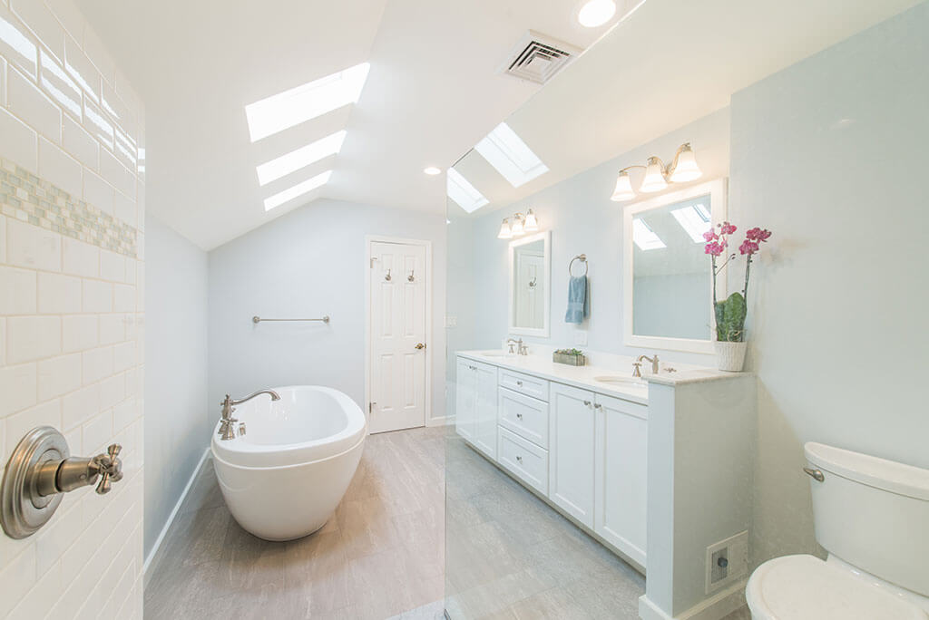 White master bathroom remodel with vaulted ceilings, skylight for natural light, his and hers vanity, white framed mirrors, clear glass shower door panel, and soaking tub in Madison, NJ renovated by JMC Home Improvement Specialists