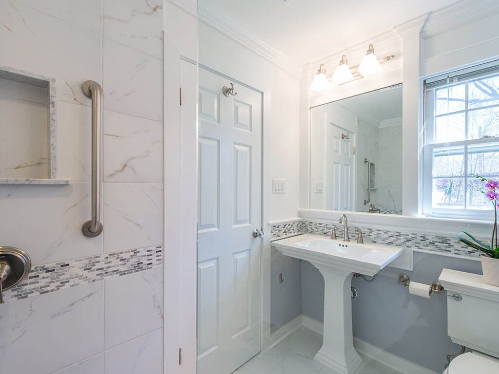 Small grey and white bathroom remodel with Kohler pedestal sink, Kohler toilet, built-in mirror and window, grey and white glass tile stripe accent and brushed nickel finishes in New Providence, NJ renovated by JMC Home Improvement Specialists