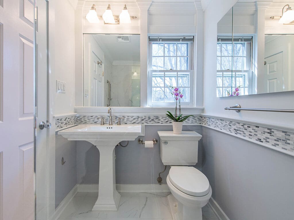 Grey and white bathroom remodel with Kohler pedestal sink, Kohler toilet, built-in mirror and window, medicine cabinet grey and white glass tile stripe accent and brushed nickel finishes in New Providence, NJ renovated by JMC Home Improvement Specialists