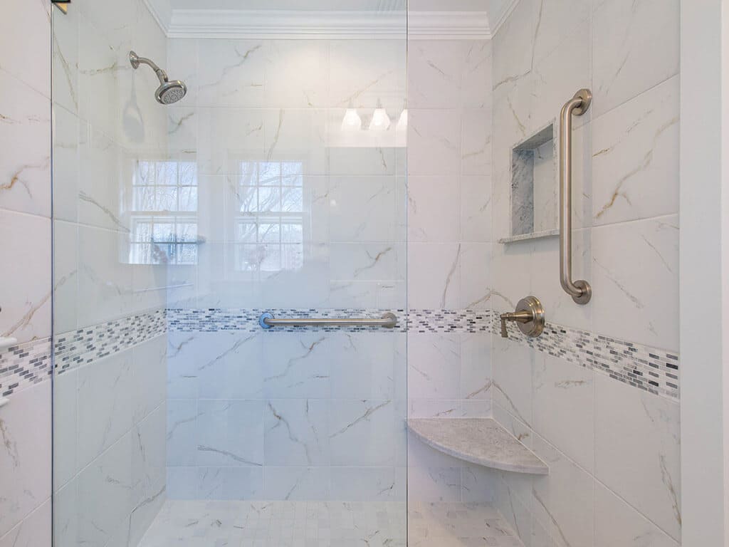 Small grey and white bathroom remodel with open shower, clear glass shower door panel, Carrera marble tile with grey and white glass tile stripe accent, niche, grab bar and brushed nickel finishes in New Providence, NJ renovated by JMC Home Improvement Specialists