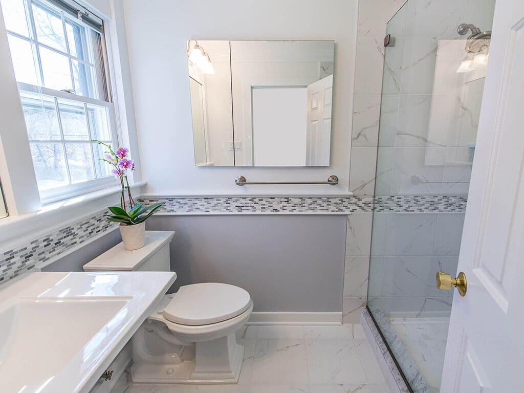Grey and white bathroom remodel with Kohler pedestal sink, Kohler toilet, medicine cabinet grey and white glass tile stripe accent and brushed nickel finishes and clear glass panel shower door in New Providence, NJ renovated by JMC Home Improvement Specialists