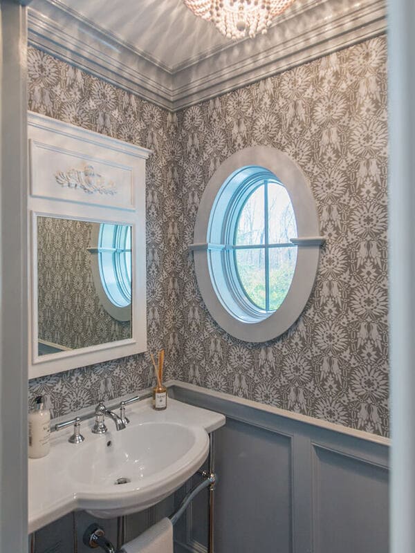 Powder room with wall panels, crown molding, retro pedestal sink with chrome legs, decorative white mirror, chandelier, Farrow & Ball paint and paper and oval window in Morris County, NJ renovated by JMC Home Improvement Specialists