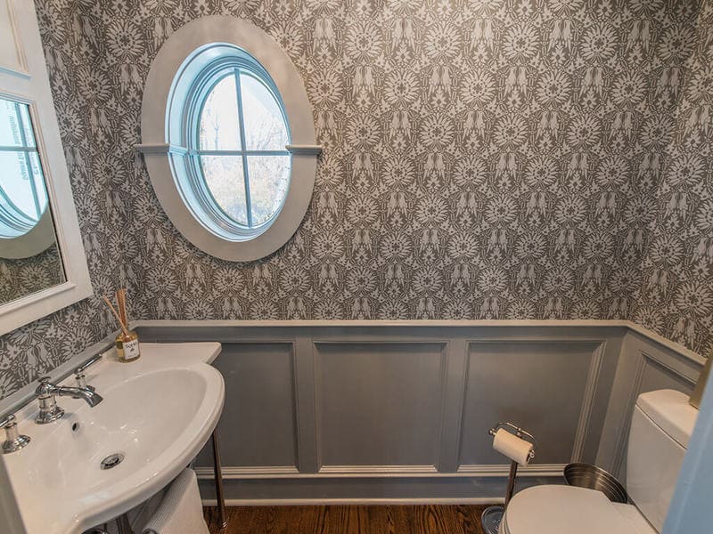 Powder room with wall panels, crown molding, retro pedestal sink with chrome legs, decorative white mirror, chandelier, Farrow & Ball paint and paper and oval window in Boonton, NJ renovated by JMC Home Improvement Specialists