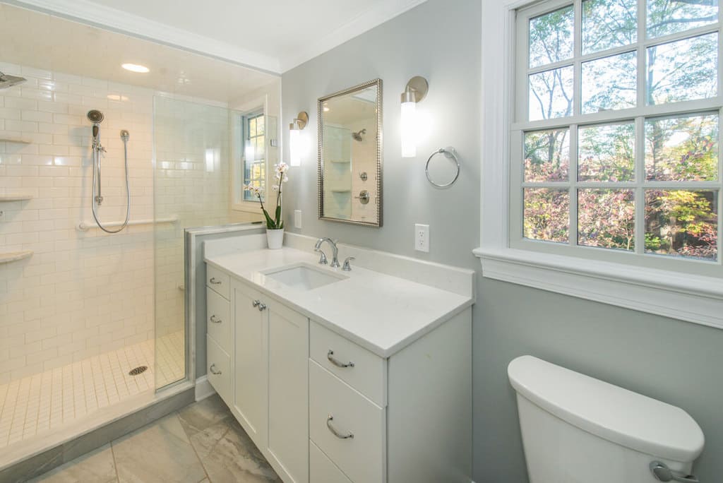 White bathroom remodel with white subway tiles in shower, marble corner shelves, grab bar, chrome finishes, white shaker cabinet vanity with quartz counter and decorative framed mirror with sconces in Springfield, NJ renovated by JMC Home Improvement Specialists