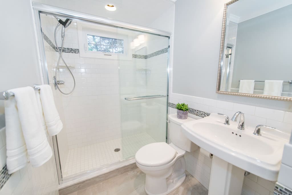 White and grey bathroom remodel with grey painted walls, white subway tiles with grey decorative glass tile strip, hand held, window in shower, chrome finishes, white pedestal sink, and decorative framed mirror in Springfield, NJ renovated by JMC Home Improvement Specialists
