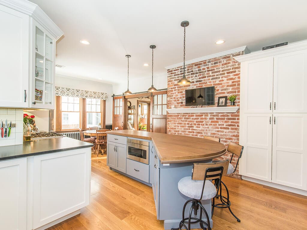 Farmhouse two tone kitchen remodel with hardwood flooring, white shaker cabinets, crown molding, subway tile backsplash, exposed brick, concrete countertop on grey island with built in microwave in Morristown, NJ renovated by JMC Home Improvement Specialists
