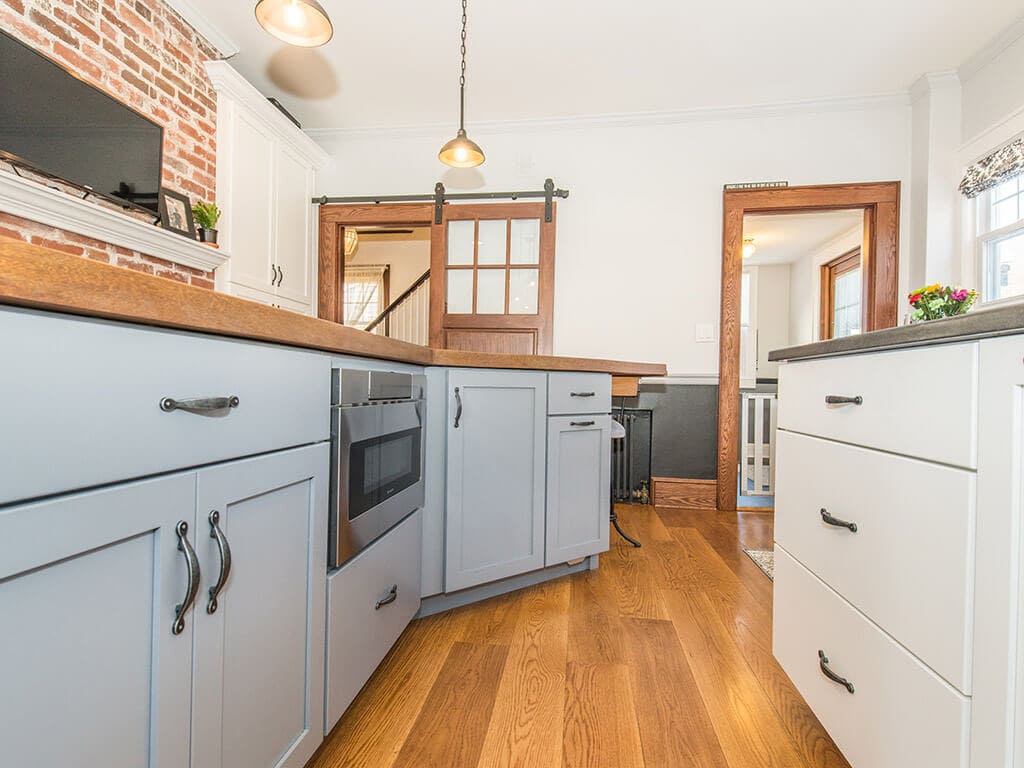 Farmhouse two tone kitchen remodel with hardwood flooring, white shaker cabinets, exposed brick, concrete countertop on grey island with built in microwave, stainless steel appliances, barn door with glass panels in Morristown, NJ renovated by JMC Home Improvement Specialists