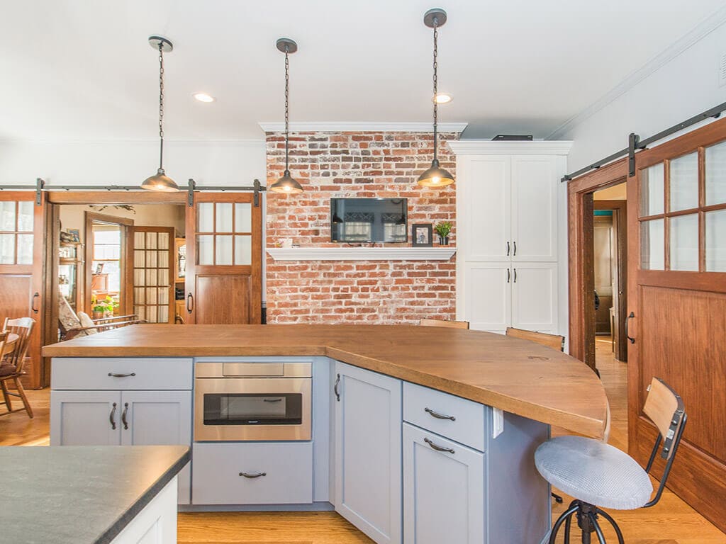 Two tone farmhouse kitchen remodel with hardwood flooring, white shaker cabinets and pantry, quartz counters and concrete countertop on grey island with built-in microwave, exposed brick, barn doors with glass panels, ss appliances in Morristown, NJ renovated by JMC Home Improvement Specialists