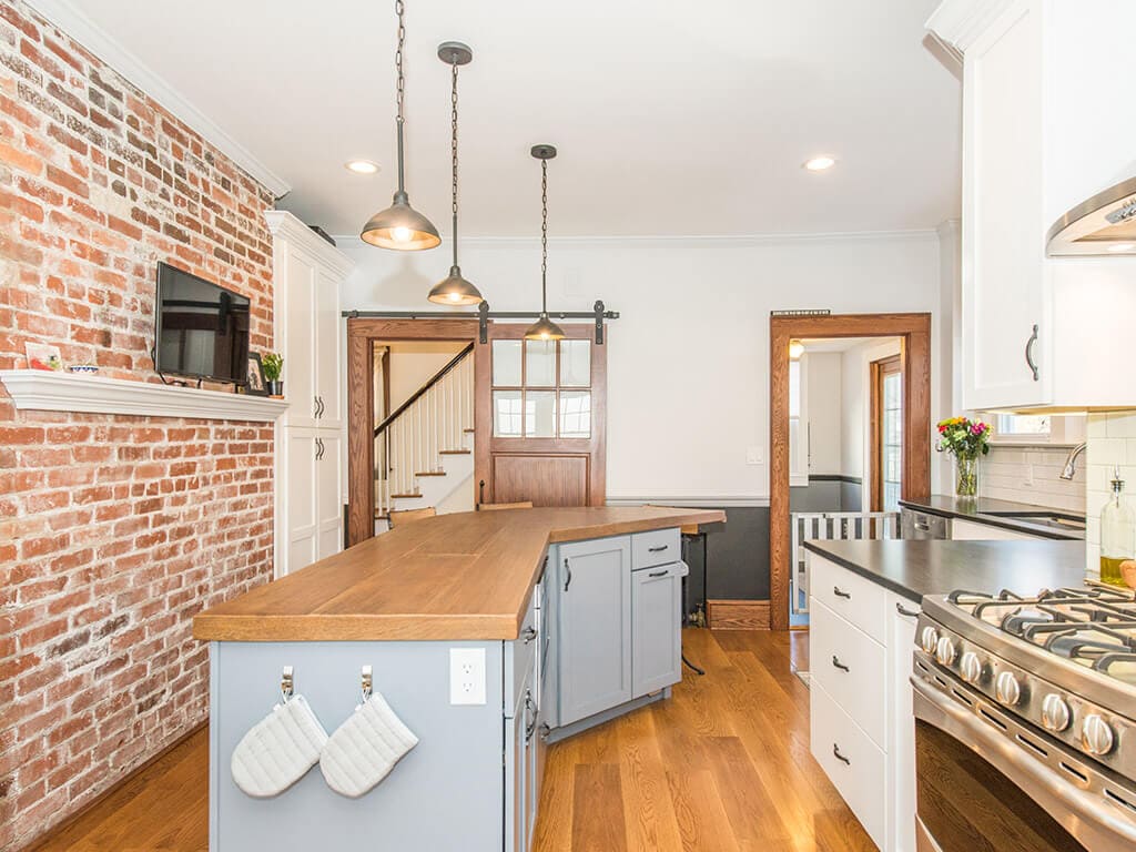 Open concept two tone farmhouse kitchen remodel with hardwood flooring, white shaker cabinet pantry, exposed brick, quartz counters and custom concrete countertop on grey island with built in microwave, barn door with glass panels, ss appliances in Morristown, NJ renovated by JMC Home Improvement Specialists