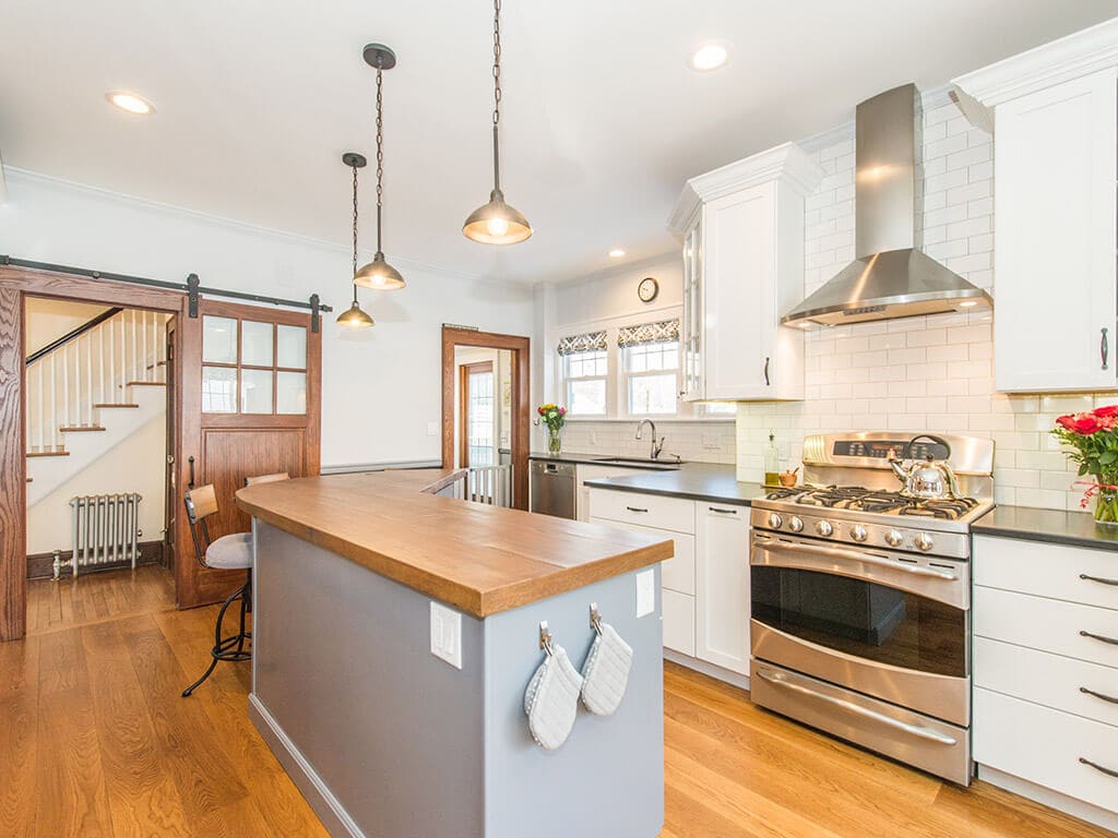 Open concept two tone farmhouse kitchen remodel with hardwood flooring, white shaker cabinet pantry, exposed brick, quartz counters and custom concrete countertop on grey island with built in microwave, barn door with glass panels,ss freestanding hood, ss appliances in Morristown, NJ renovated by JMC Home Improvement Specialists