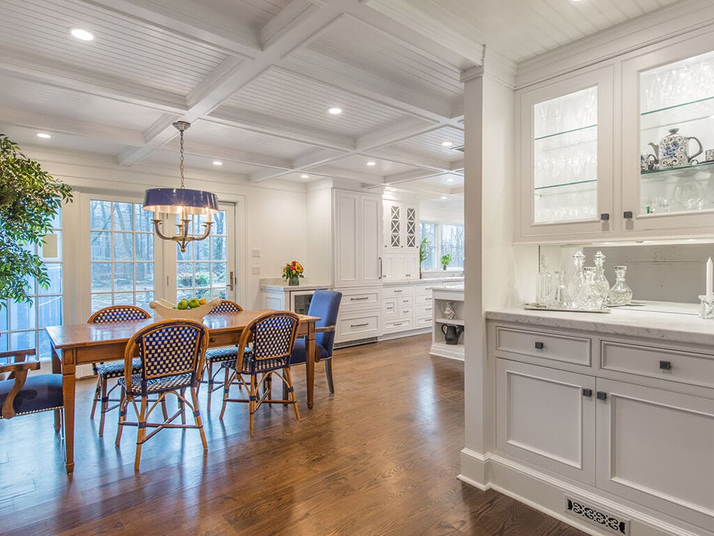 Award winning open concept cape cod kitchen with bar area with glass cabinets and lighting, white inset shaker cabinets with marble counter, dining area, coffered ceiling and 7” oak flooring throughout in Boonton, NJ renovated by JMC Home Improvement Specialists