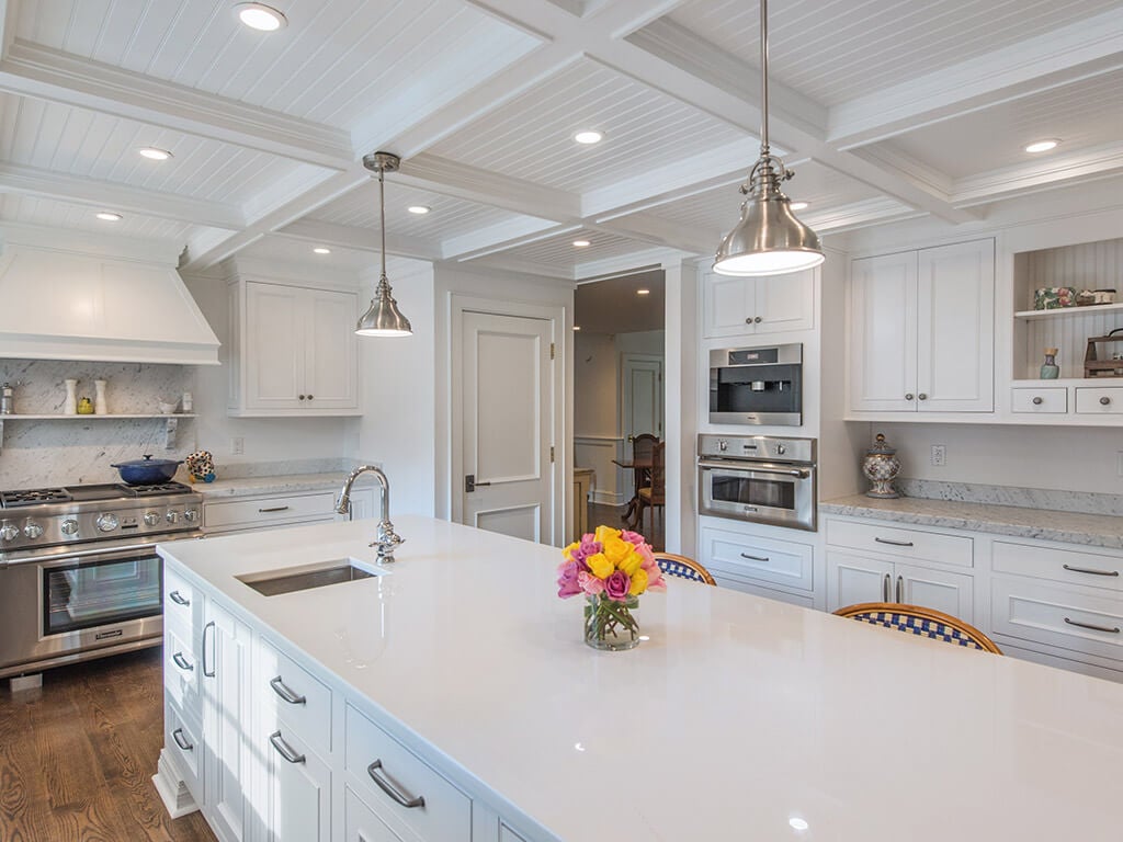 Cape cod white kitchen remodel with white inset shaker cabinets, nano glass counter at island with sink, 2” thick carerra marble counters, built in coffee machine, open shelving with wainscot  panels to match ceiling panels, custom wood hood over range with 7” oak hardwood flooring in Boonton, NJ renovated by JMC Home Improvement Specialists