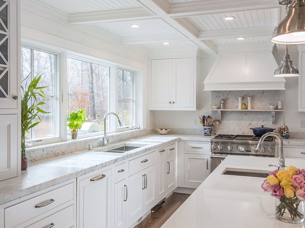 Farmhouse white kitchen remodel with white inset shaker cabinets, nano glass counter at island with sink, 2” thick carerra marble counters, 4” splash, marble behind stove  with shelf, coffered ceiling with wainscot ceiling panels and LED highhats, triple window above sink in Boonton, NJ renovated by JMC Home Improvement Specialists