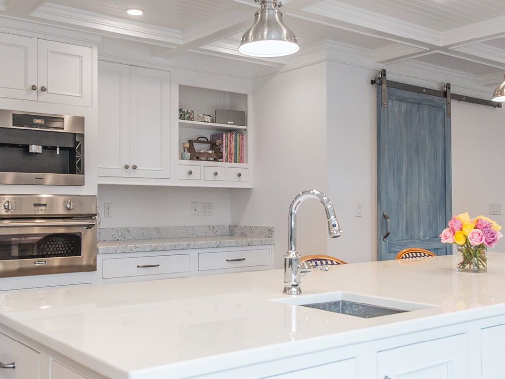 Award winning Farmhouse Cape cod white kitchen remodel with white inset shaker cabinets, nano glass counter at island with sink, 2” thick carerra marble counters, built in coffee machine, bookshelf with wainscot panels to match ceiling panels, blue barn pantry door in Boonton, NJ renovated by JMC Home Improvement Specialists