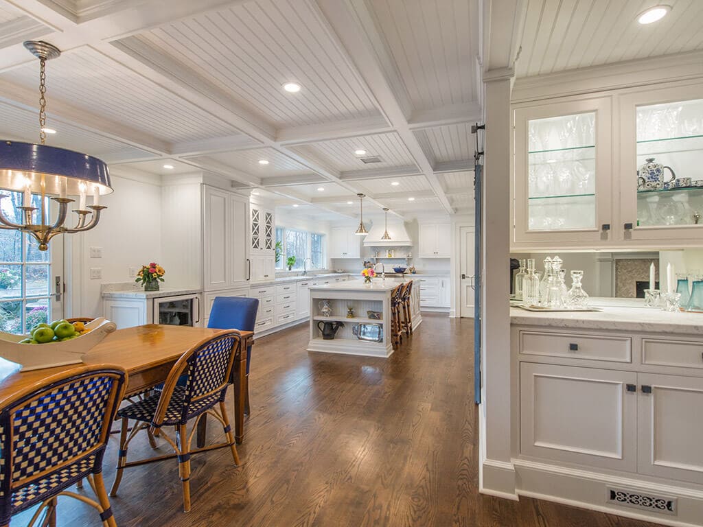 Award winning white farmhouse open concept cape cod kitchen with bar area with glass cabinets and lighting, white inset shaker cabinets with marble counter, dining area, coffered ceiling, blue accents and 7” oak flooring throughout in Boonton, NJ renovated by JMC Home Improvement Specialists