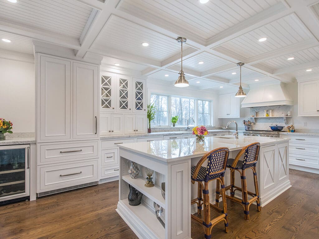 White farmhouse open concept cape cod kitchen with inset shaker cabinets, built-in refrigerator, beverage fridge, marble counter, nano glass counter on island with open shelving and seating, coffered ceiling, highhats and pendant lighting, custom wood hood and 7” oak hardwood flooring throughout in Boonton, NJ renovated by JMC Home Improvement Specialists