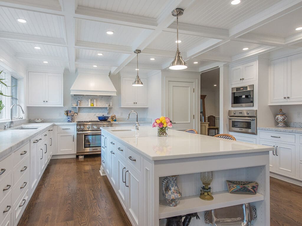 White farmhouse open concept cape cod kitchen with inset shaker cabinets, built-in refrigerator, beverage fridge, marble counter, nano glass counter on island with open shelving and seating, coffered ceiling, highhats and pendant lighting, custom wood hood and 7” oak hardwood flooring throughout in Boonton, NJ renovated by JMC Home Improvement Specialists