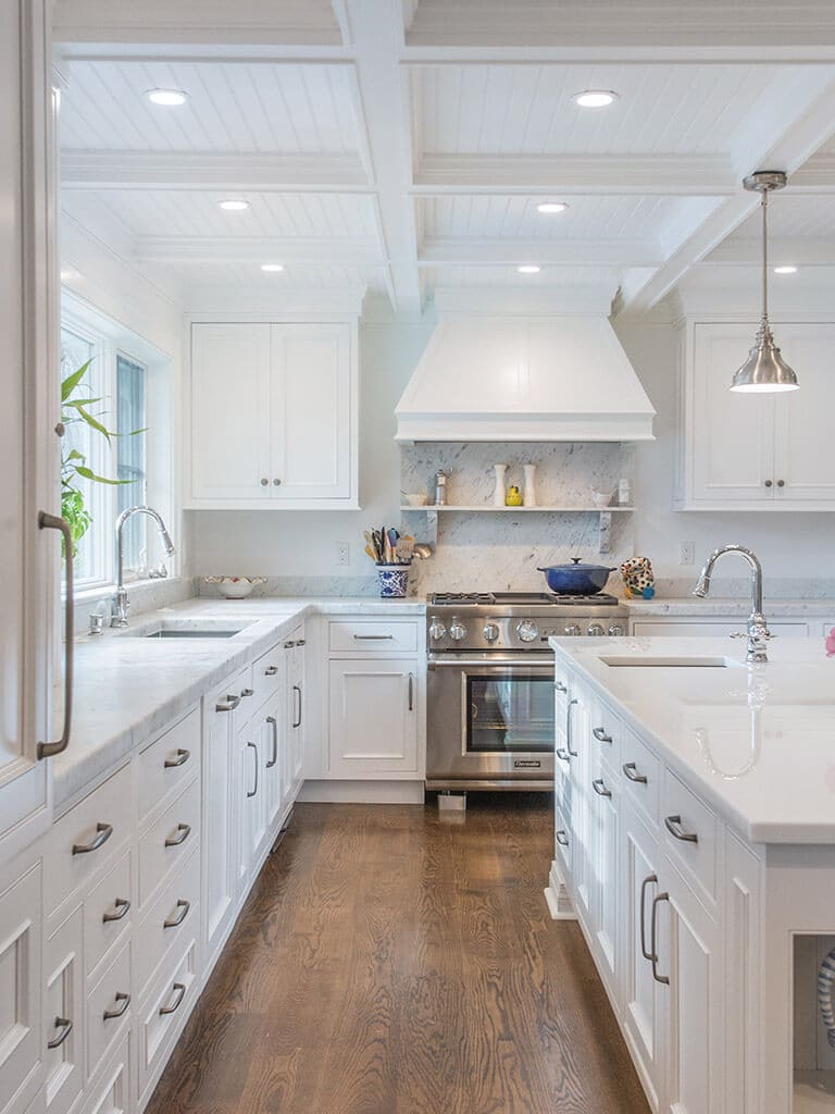 Open concept white cape cod farmhouse kitchen with inset shaker cabinets, marble counter, nano glass counter on island with open shelving and seating, two sinks, coffered ceiling, highhats and pendant lighting, custom wood hood and 7” oak hardwood flooring throughout in Boonton, NJ renovated by JMC Home Improvement Specialists