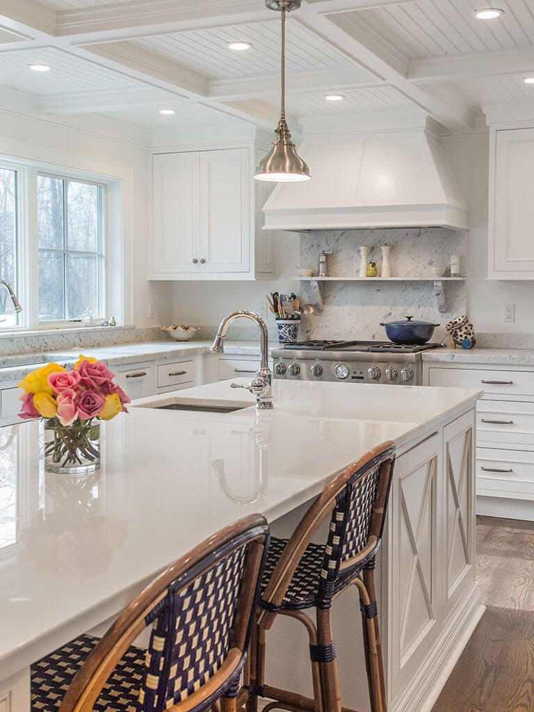 White cape cod farmhouse kitchen with inset shaker cabinets, marble counter, nano glass counter on island with open shelving and seating, two sinks, coffered ceiling, highhats and pendant lighting, custom wood hood and 7” oak hardwood flooring throughout in Boonton, NJ renovated by JMC Home Improvement Specialists