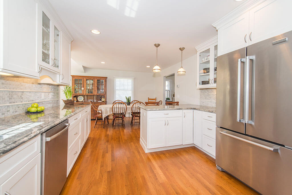 Open concept white kitchen remodel with white shaker cabinets, glass upper cabinet, granite countertops, pendant lighting, stainless appliances, dining area with hardwood flooring in Chatham, NJ renovated by JMC Home Improvement Specialists