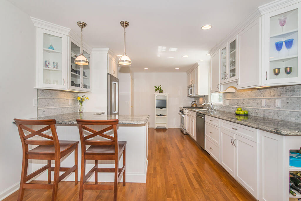 Open concept white kitchen remodel with white shaker cabinets, glass upper cabinet, granite countertops, pendant lighting, highhats, stainless appliances, seating at peninsula with hardwood flooring in Chatham, NJ renovated by JMC Home Improvement Specialists