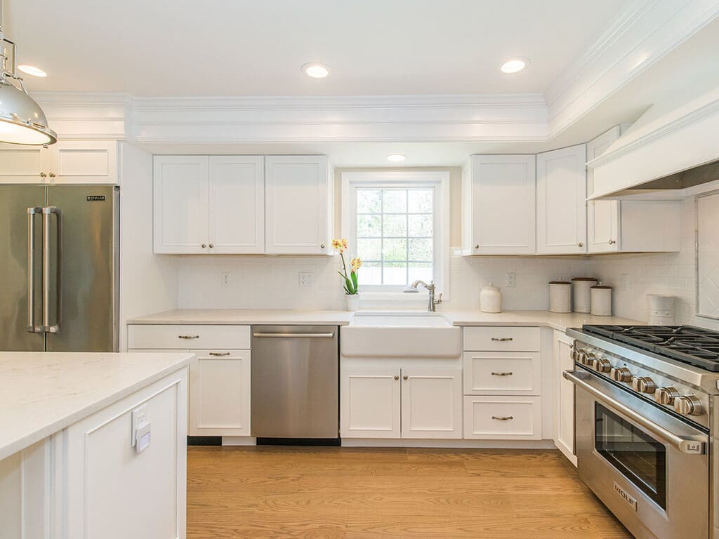 Open concept white kitchen remodel with white pantry cabinets, glass upper cabinet doors, quartz countertops, crown molding, pendant lighting over island and hardwood flooring in Morristown, NJ renovated by JMC Home Improvement Specialists