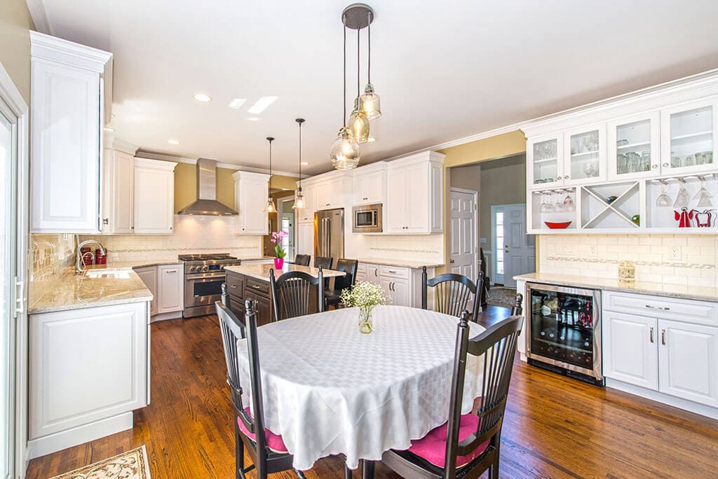 Open concept two tone kitchen remodel with cherry island and white cabinets, crown molding, granite counters, pendants over island, subway tile backsplash with mosaic strip, stainless appliances, custom bar and hardwood floor throughout in Morris County, NJ renovated by JMC Home Improvement Specialists