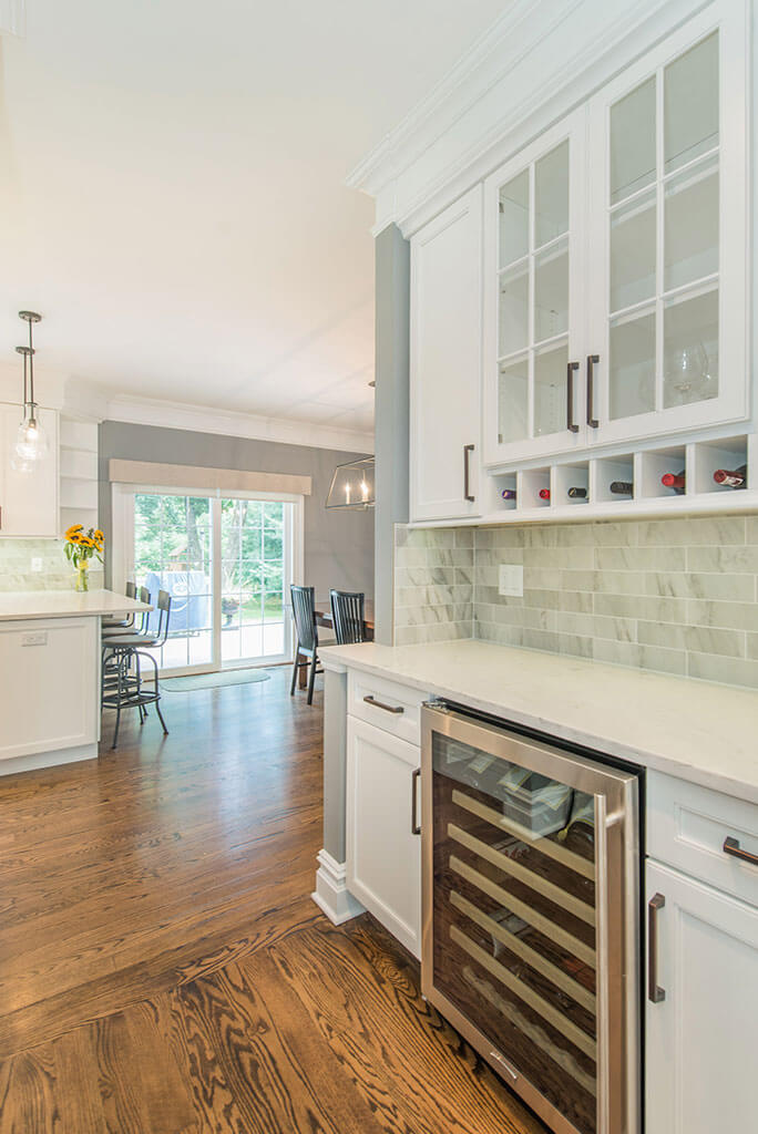 White open floor plan kitchen remodel with shaker pantry cabinets, with built in wine rack, glass cabinet doors, wine fridge, quartz counters, and marble backsplash in Florham Park, NJ renovated by JMC Home Improvement Specialists