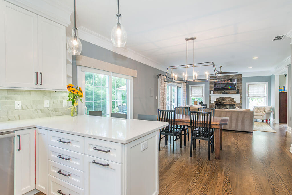 Open concept white kitchen remodel with shaker cabinets, quartz counters, pendants over peninsula, marble backsplash, dining area and hardwood floors throughout in Florham Park, NJ renovated by JMC Home Improvement Specialists