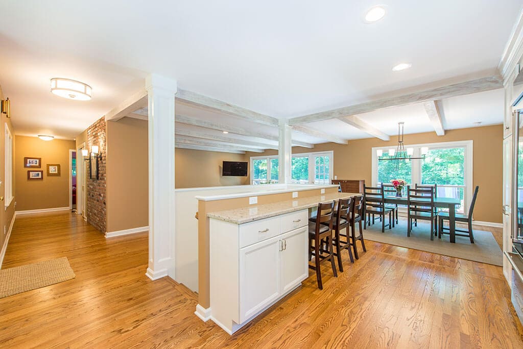 Open floor plan white kitchen remodel with shaker cabinets, quartz counters, wood beams, exposed brick in  hall and hardwood flooring throughout in Mendham, NJ renovated by JMC Home Improvement Specialists