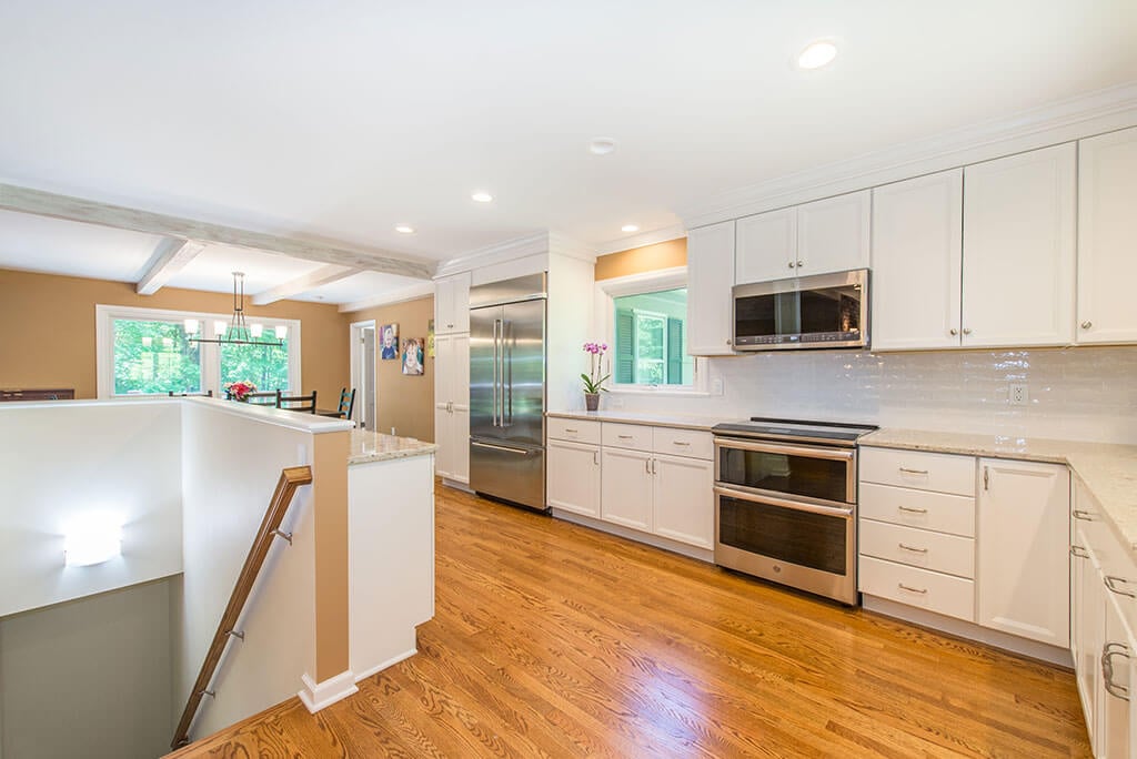 Open floor plan white kitchen remodel with shaker cabinets, quartz counters, stainless appliances, wood beams, stairs to basement and hardwood flooring throughout in Mendham, NJ renovated by JMC Home Improvement Specialists