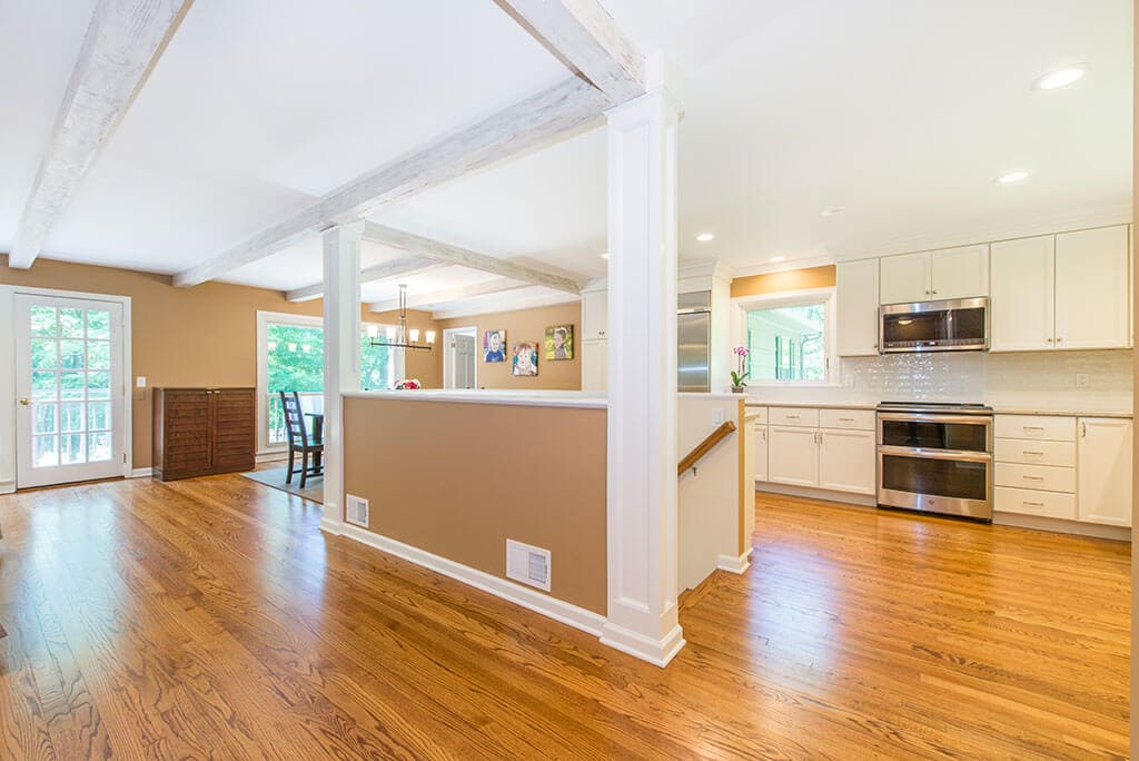 Open floor plan white kitchen remodel with shaker cabinets, quartz counters, stainless appliances, wood beams, stairs to basement, dining area and hardwood flooring throughout in Mendham, NJ renovated by JMC Home Improvement Specialists