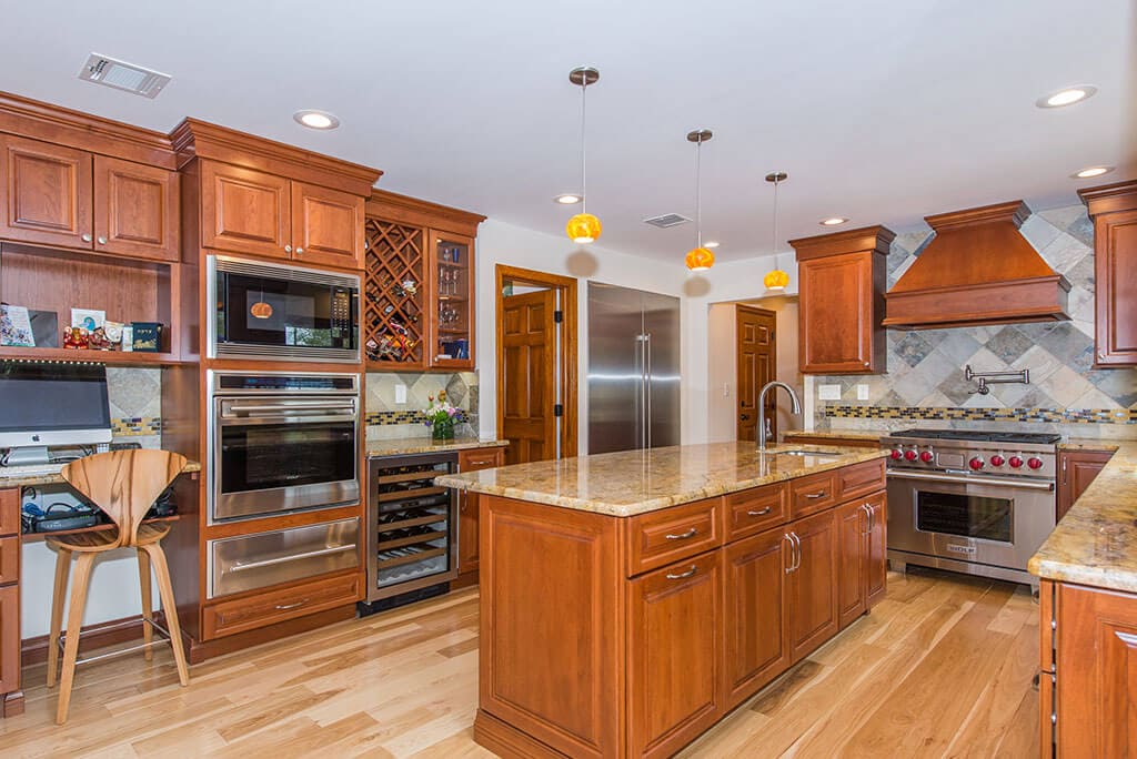 Wood cabinetry in kitchen remodel with freestanding wood hood, granite counters, built in ovens, wine fridge, desk area, build in shelving, crown molding, pendants over island, pot filler with mosaic backsplash in Morristown, NJ remodeled by JMC Home Improvement Specialists