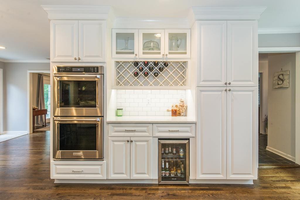 Double oven, glass cabinet doors above wine rack, bar fridge, subway tile backsplash, white cabinets with undercabinet lighting, tall pantry cabinets with hardwood flooring in Denville, NJ remodeled by JMC Home Improvement Specialists