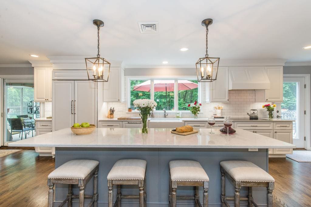 Two tone open concept kitchen with grey island with quartz counter, bar stools, panel refrigerator, three panel windows above apron sink, wood hood over cooktop, subway tile backsplash with undercabinet lighting in Denville, NJ remodeled by JMC Home Improvement Specialists