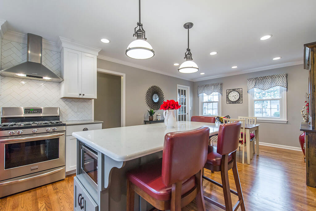 Eat in two tone open concept kitchen remodel with herringbone white backsplash, freestanding hood, white shaker cabinets, quartz counters, pendant lighting over island and LED highhats, grey and white cabinets with red accents in Parsippany, NJ remodeled by JMC Home Improvement Specialists